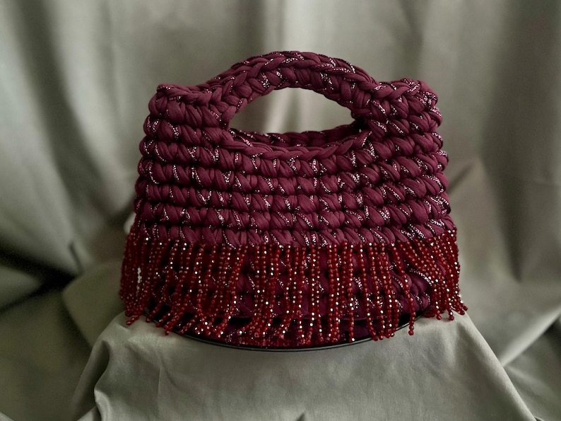 May include: A burgundy crocheted handbag with a rounded handle and beaded fringe. The bag is made of a thick, textured yarn with silver metallic threads woven throughout. The fringe is made of small, dark red beads.