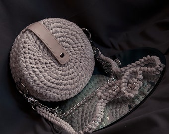 May include: A round, gray crocheted bag with a leather strap and a silver chain strap. The bag is reflected in a mirror.