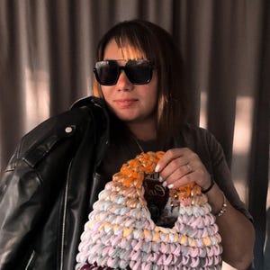 May include: A colorful, handmade crochet bag with a unique design. The bag features a mix of pastel colors, including pink, orange, and purple. A black leather jacket is held in the background.