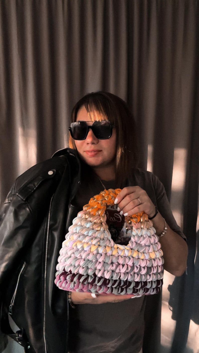 May include: A colorful, handmade crochet bag with a unique design. The bag features a mix of pastel colors, including pink, orange, and purple. A black leather jacket is held in the background.