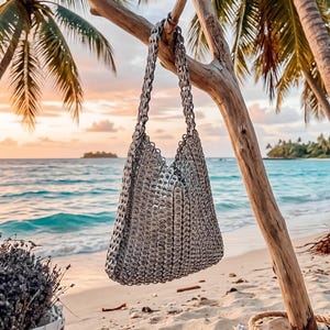 May include: A silver tote bag made from recycled materials hangs from a tree branch on a beach. The bag has a chain strap and a woven design. The background features a tropical beach scene with palm trees, sand, and ocean.