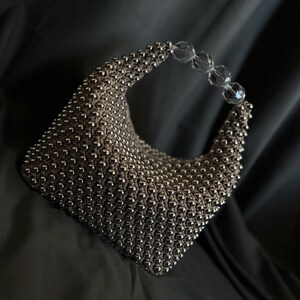 May include: A silver beaded handbag with a clear plastic handle. The bag is made of small, round beads that are woven together to create a textured surface.