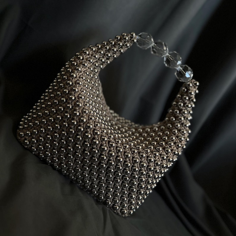 May include: A silver beaded handbag with a clear plastic handle. The bag is made of small, round beads that are woven together to create a textured surface.