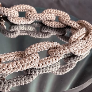 May include: A close-up of a beige crocheted chain link bag strap. The strap is made of thick yarn and has a chunky, textured look.