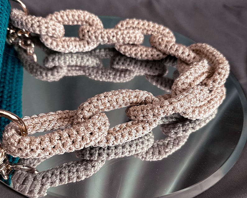 May include: A close-up of a beige crocheted chain link bag strap. The strap is made of thick yarn and has a chunky, textured look.