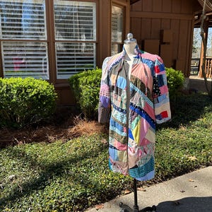 May include: A patchwork coat in various colors, including pink, blue, and green, is displayed on a black metal stand. The coat features a variety of fabric patterns and textures, creating a unique and artistic design. The coat is displayed outdoors.