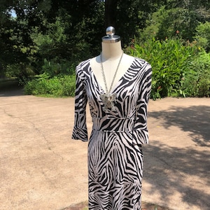 May include: A black and white zebra print dress with a v-neckline and 3/4 sleeves. The dress is on a mannequin and has a silver necklace with a large pendant.