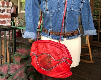 May include: A red leather purse with a silver zipper and a decorative design. The purse is being worn on a mannequin with a denim jacket and a brown belt.