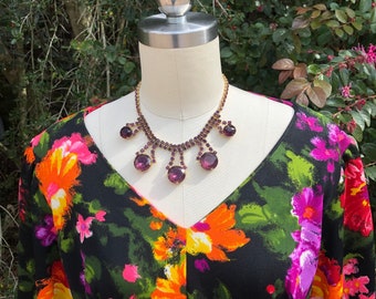 May include: A black dress with a floral print featuring pink, orange, and green flowers. The dress has a V-neckline and long sleeves. A purple rhinestone necklace with dangling stones is worn on a mannequin.