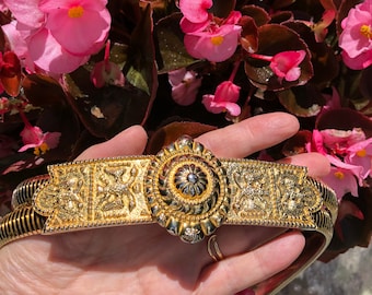 Könnte beinhalten: Ein goldfarbener Gürtel mit einer verzierten, runden Schnalle. Die Schnalle hat ein erhabenes, strukturiertes Design und eine kleine, silberfarbene Mitte.