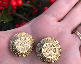 May include: A pair of gold-tone earrings with a circular design. The earrings feature a raised, white enamel logo that reads "Paris New York Givenchy".