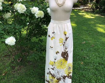 May include: A cream-colored, floor-length skirt with a floral and butterfly design. The skirt features large yellow, white, and brown flowers, with small black and yellow butterflies scattered across the fabric. A multi-strand pearl necklace is draped on a mannequin.