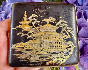 May include: A black cigarette case with a gold design of a Japanese pagoda and trees. The case is decorated with a gold-colored design of a pagoda and trees.