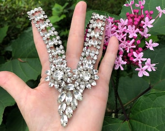 May include: A silver tone rhinestone necklace with a V-shaped design. The necklace features a large cluster of rhinestones at the center and smaller clusters along the sides.