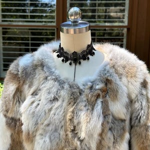 May include: A fur coat in a mix of brown, white, and grey hues, presented on a mannequin. A black lace choker with hanging beads is around the mannequin's neck. The coat has a soft, fluffy texture, implying warmth and sophistication.