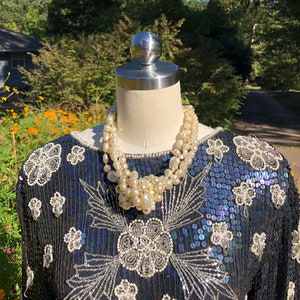 Puede incluir: Una blusa negra de lentejuelas con bordados plateados y un collar de perlas. La blusa tiene un escote redondo y mangas largas.