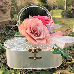 Könnte beinhalten: Eine weiße, transparente, rechteckige Box aus Kunststoff mit einem goldenen Verschluss. Die Box hat einen transparenten Kunststoffgriff und ist mit einer rosa und weißen Rose verziert.