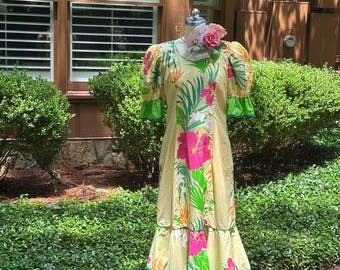 Könnte beinhalten: Ein bodenlanges gelbes Kleid mit einem lebendigen tropischen Druck aus rosa Hibiskusblüten und grünen Palmblättern. Das Kleid hat kurze Puffärmel, einen Rundhalsausschnitt und einen gestuften, gerüschten Saum mit hellgrüner Kante.
