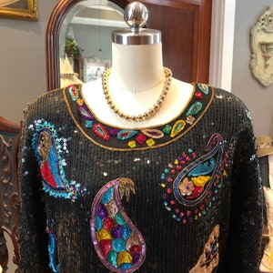 Puede incluir: Una blusa negra de lentejuelas con bordados de paisley coloridos y un collar de cadena dorada. La blusa tiene un escote redondo y mangas largas.