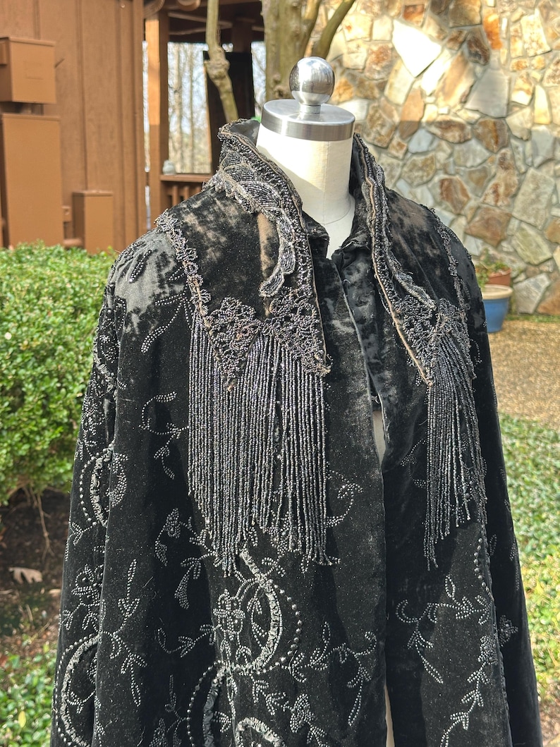 May include: Black velvet cape with intricate silver embroidery and beaded fringe detailing. The cape features a high collar and a flowing design, showcasing a vintage aesthetic. The embroidery includes floral and swirling patterns.