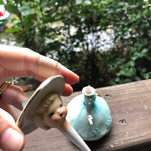 Może przedstawiać: Vintage kork do butelki wykonany z porcelany w kształcie lalki w niebieskiej sukience i białym kapeluszu. Głowa lalki jest oddzielona od ciała i jest trzymana w dłoni.
