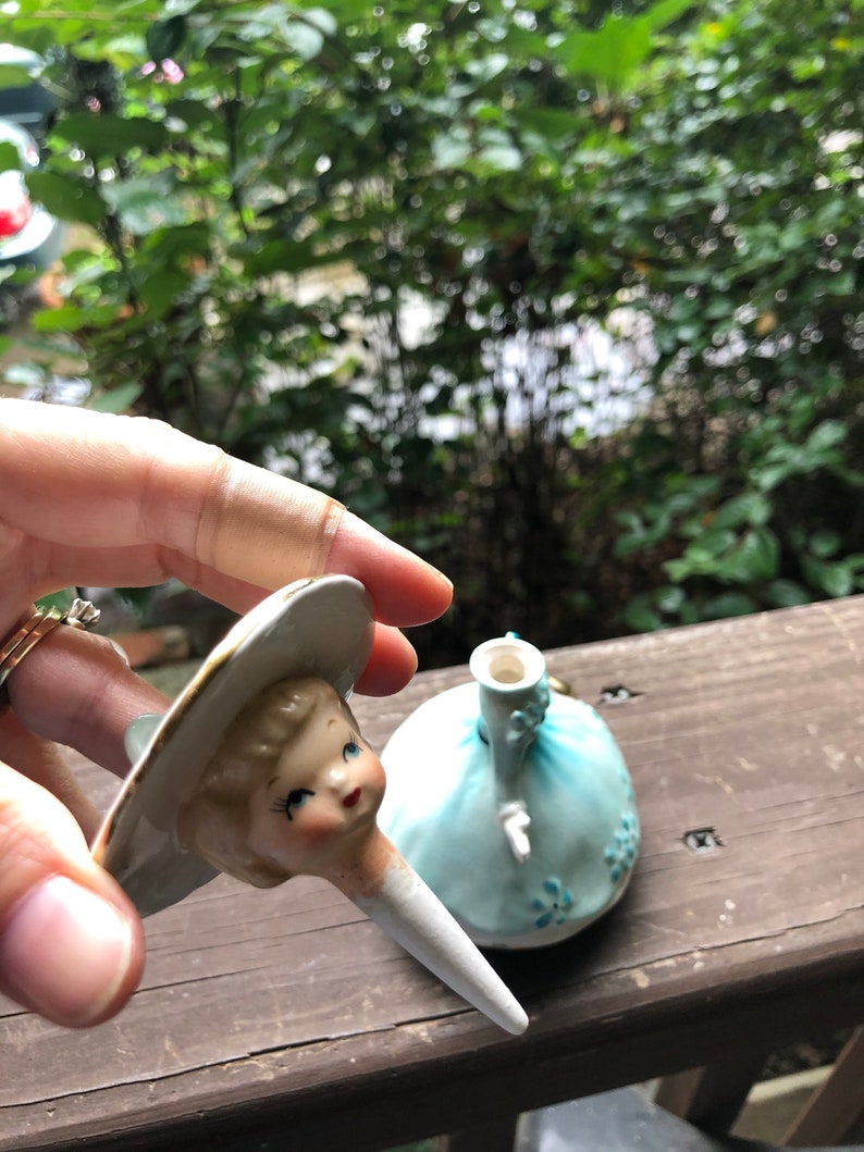 Może przedstawiać: Vintage kork do butelki wykonany z porcelany w kształcie lalki w niebieskiej sukience i białym kapeluszu. Głowa lalki jest oddzielona od ciała i jest trzymana w dłoni.