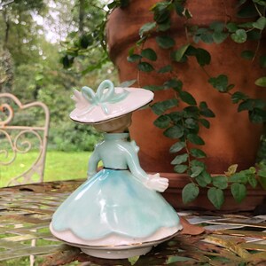 Może przedstawiać: Porcelanowa figurka kobiety w jasnoniebieskiej sukience i białym kapeluszu z zieloną kokardką. Figurka stoi na metalowym stole z zieloną rośliną w tle.