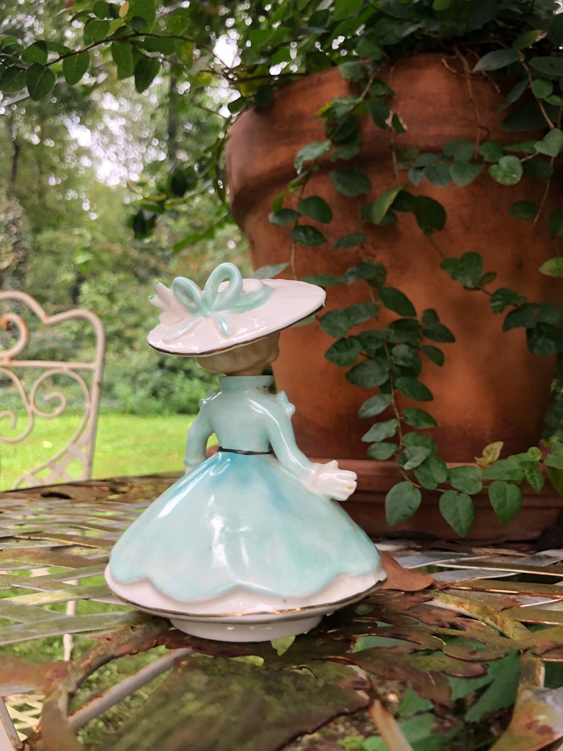 Może przedstawiać: Porcelanowa figurka kobiety w jasnoniebieskiej sukience i białym kapeluszu z zieloną kokardką. Figurka stoi na metalowym stole z zieloną rośliną w tle.