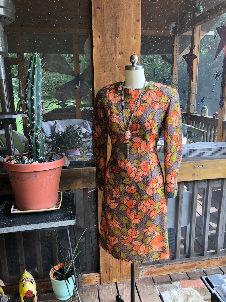 Peut inclure: Une robe vintage &agrave; imprim&eacute; floral dans les tons orange, vert et marron. La robe a des manches longues et une silhouette ajust&eacute;e.