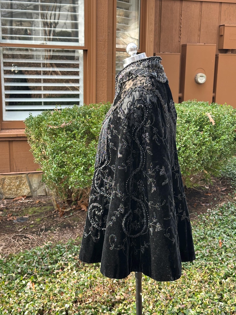 May include: Black velvet cape with intricate silver beadwork and embroidery. The cape features a high collar and a flared silhouette, with detailed floral and scroll patterns. The garment is displayed on a mannequin.