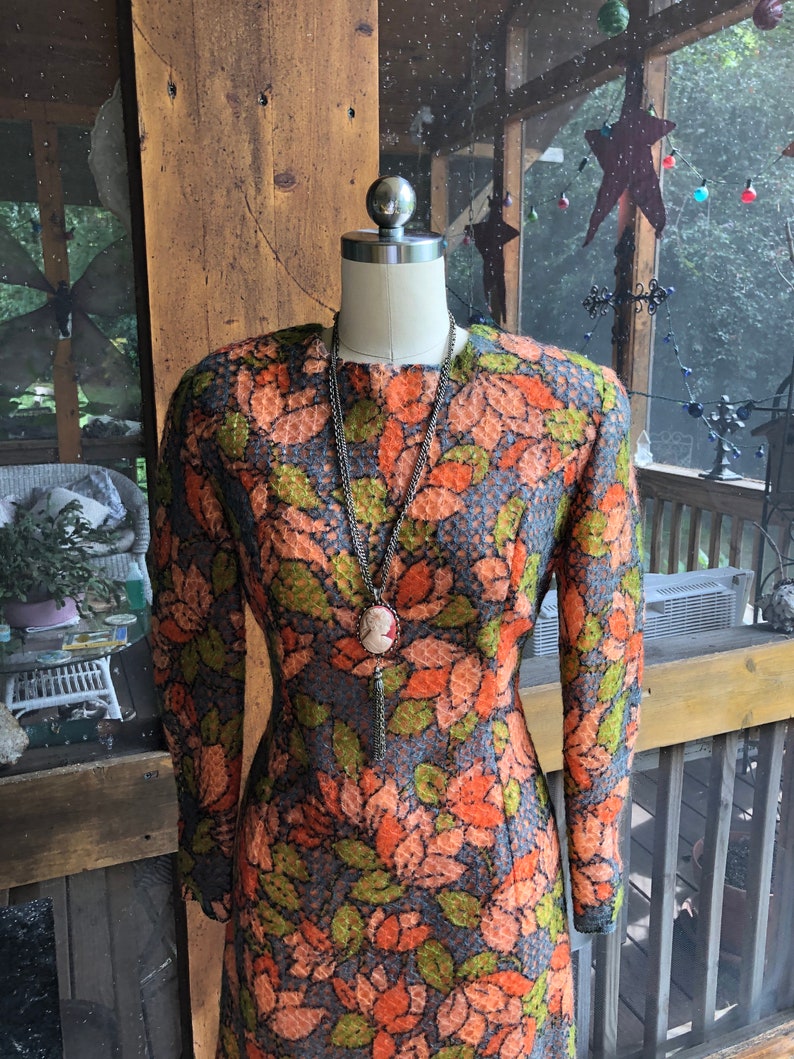 Peut inclure: Une robe vintage &agrave; motif floral dans les tons orange, vert et bleu. La robe a des manches longues et une silhouette ajust&eacute;e. Elle est expos&eacute;e sur un mannequin.