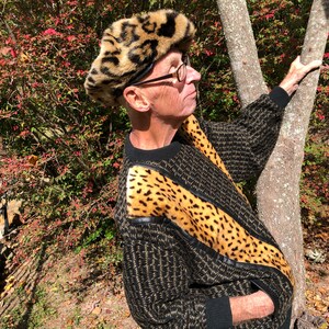 May include: A man wearing a black and brown sweater with a leopard print trim and a leopard print fur hat. He is standing in front of a tree.