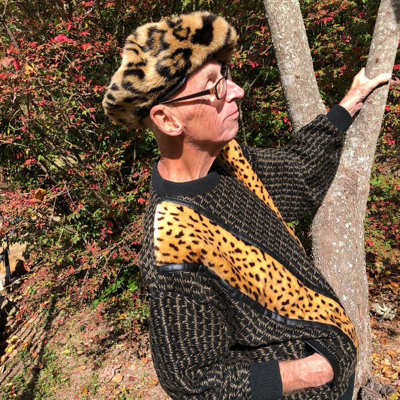 May include: A man wearing a black and brown sweater with a leopard print trim and a leopard print fur hat. He is standing in front of a tree.