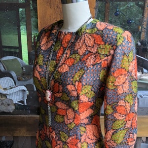 Peut inclure: Une robe vintage &agrave; motif floral dans les tons orange, vert et bleu. La robe a des manches longues et une silhouette ajust&eacute;e. Elle est expos&eacute;e sur un mannequin avec un collier en cha&icirc;ne argent&eacute;e.
