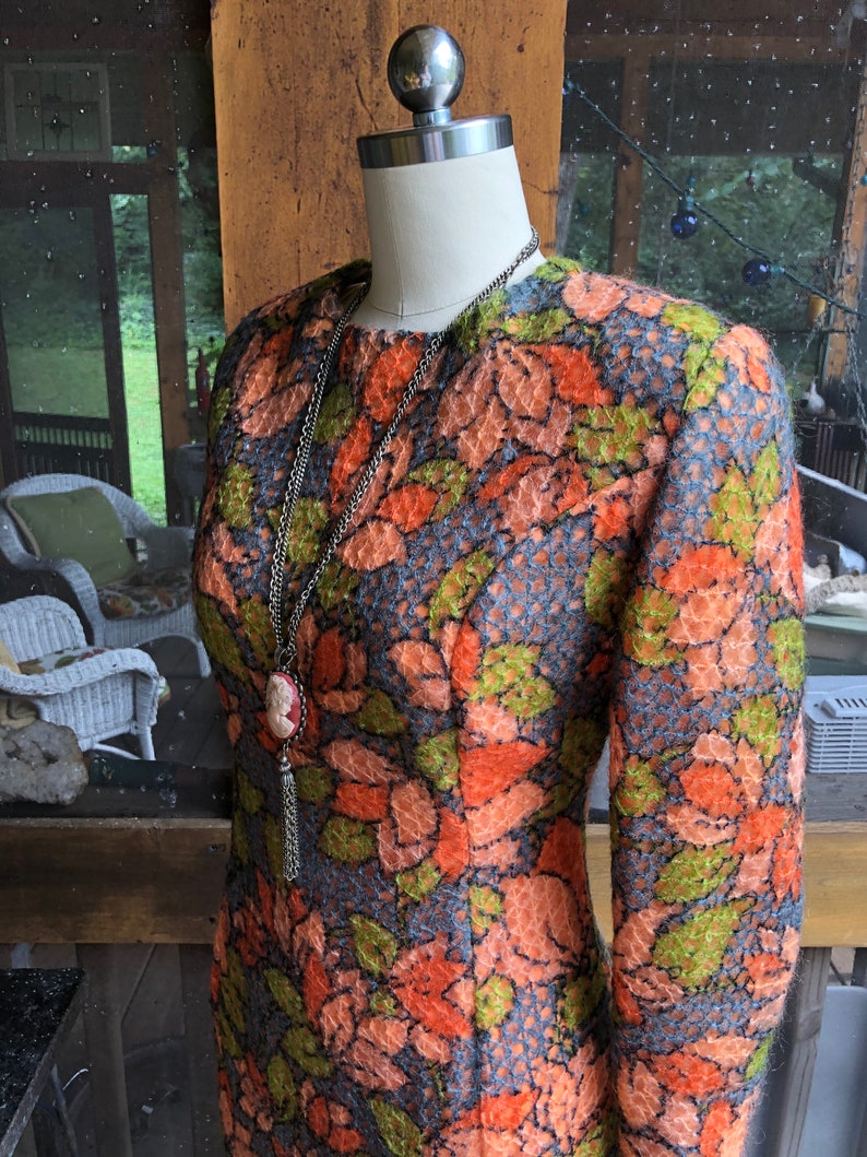 Peut inclure: Une robe vintage &agrave; motif floral dans les tons orange, vert et bleu. La robe a des manches longues et une silhouette ajust&eacute;e. Elle est expos&eacute;e sur un mannequin avec un collier en cha&icirc;ne argent&eacute;e.