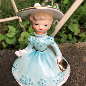 Może przedstawiać: Porcelanowa figurka dziewczyny w niebieskiej sukience i białym kapeluszu z złotą obw&oacute;dką. Trzyma złotą torebkę i ma niebieskie kwiaty na sukience.