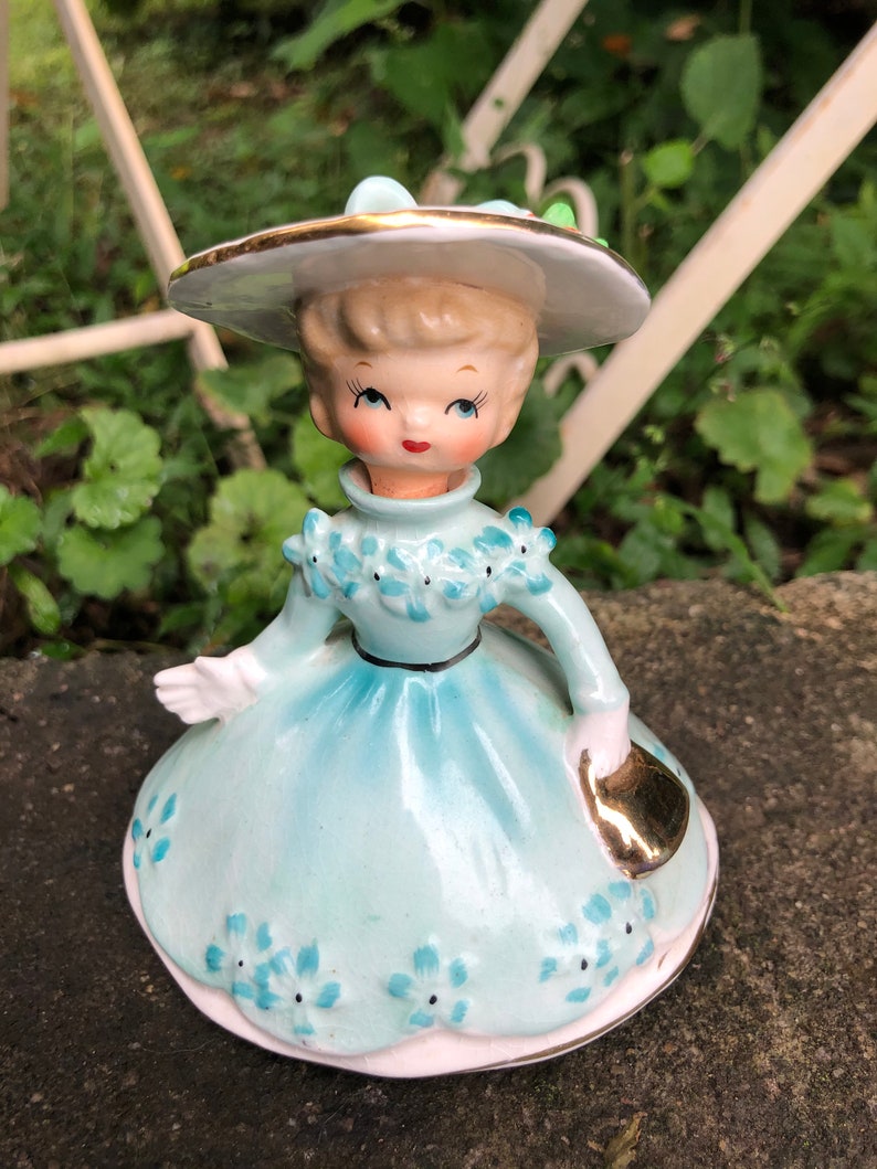 Może przedstawiać: Porcelanowa figurka dziewczyny w niebieskiej sukience i białym kapeluszu z złotą obw&oacute;dką. Trzyma złotą torebkę i ma niebieskie kwiaty na sukience.