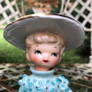 Może przedstawiać: Porcelanowa figurka przedstawiająca dziewczynkę w niebieskiej sukience z białymi kwiatami i białym kapeluszu z złotą obw&oacute;dką. Figurka ma namalowaną twarz z niebieskimi oczami i czerwonymi ustami.