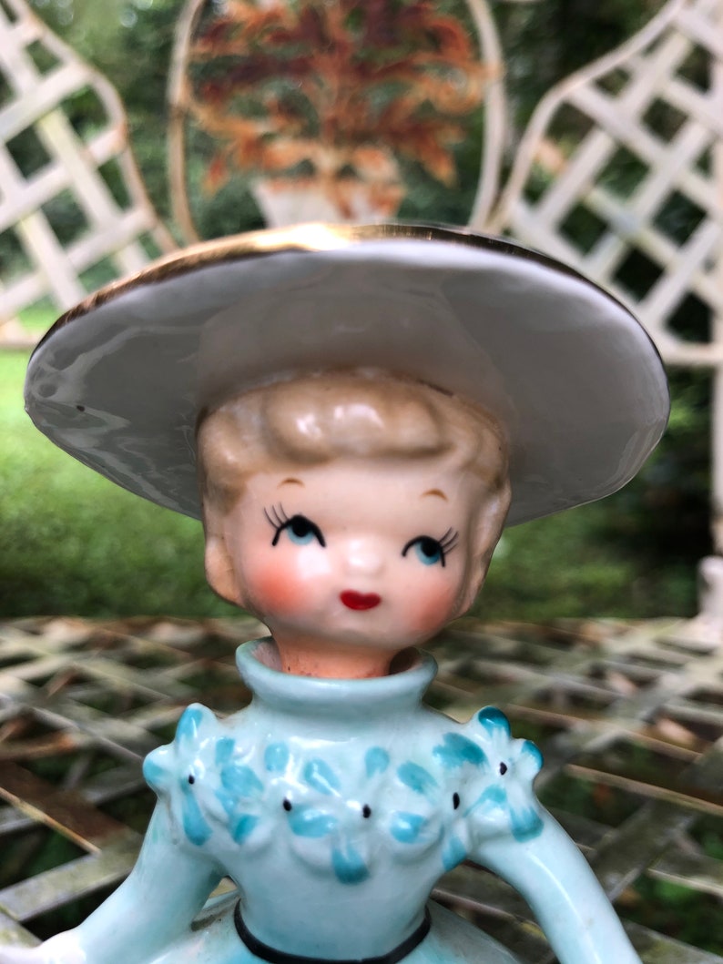 Może przedstawiać: Porcelanowa figurka przedstawiająca dziewczynkę w niebieskiej sukience z białymi kwiatami i białym kapeluszu z złotą obw&oacute;dką. Figurka ma namalowaną twarz z niebieskimi oczami i czerwonymi ustami.
