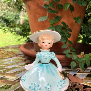 Może przedstawiać: Winietkowa figurka porcelanowa przedstawiająca dziewczynkę w niebieskiej sukience z białymi kwiatami i białym kapeluszu z złotą ozdobą. Dziewczynka trzyma złotą torebkę.