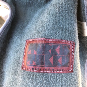 May include: Close-up of a brown leather patch with white stitching. The patch has the text "MUKAVA" in a stylized font and the text "MADE IN FINLAND" below it.