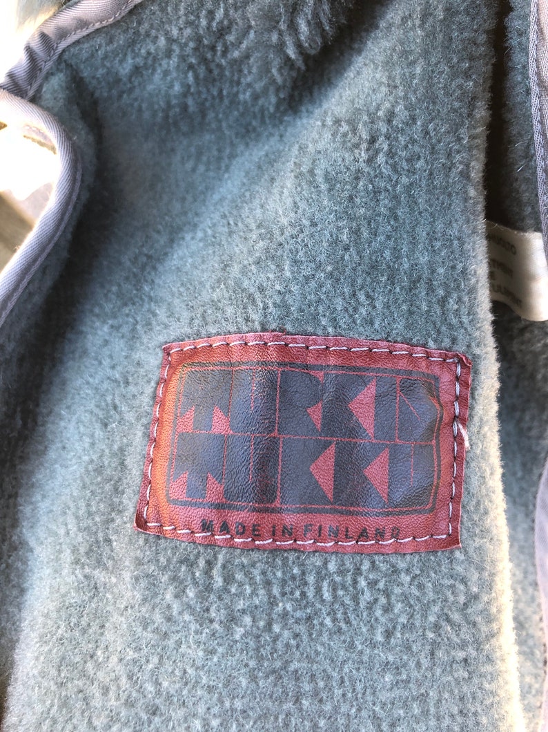 May include: Close-up of a brown leather patch with white stitching. The patch has the text "MUKAVA" in a stylized font and the text "MADE IN FINLAND" below it.
