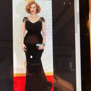 Puede incluir: Una mujer con un vestido negro de manga larga, largo hasta el suelo, con detalles de encaje en los hombros y el escote. Est&aacute; de pie sobre una alfombra roja con un escenario de fondo.