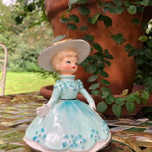 Może przedstawiać: Porcelanowa figurka przedstawiająca dziewczynkę w jasnoniebieskiej sukience, białym kapeluszu z złotą ozdobą. Sukienka jest ozdobiona niebieskimi kwiatami. Figurka stoi na metalowym stole.