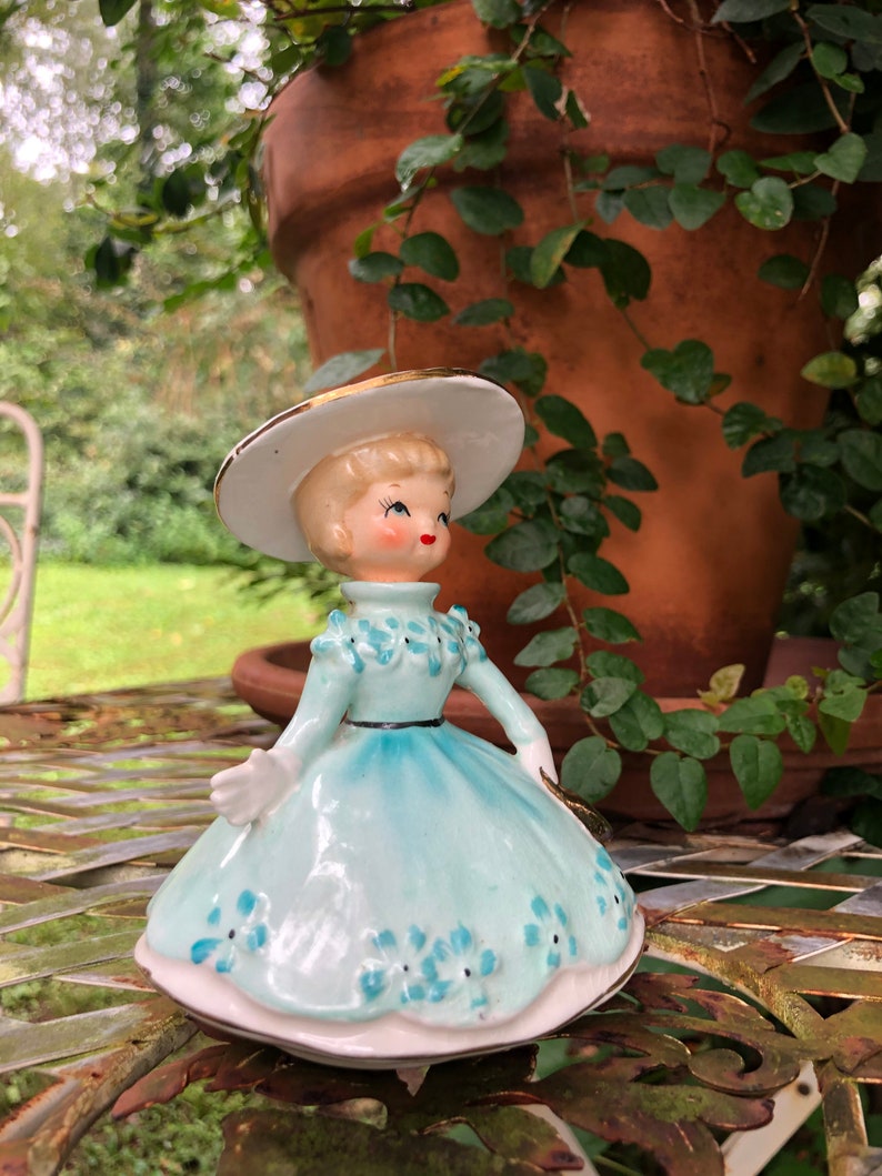 Może przedstawiać: Porcelanowa figurka przedstawiająca dziewczynkę w jasnoniebieskiej sukience, białym kapeluszu z złotą ozdobą. Sukienka jest ozdobiona niebieskimi kwiatami. Figurka stoi na metalowym stole.