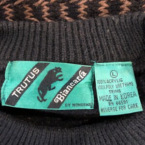 May include: A black and brown sweater with a geometric pattern. The sweater has a green tag with the text "TRUTUS Biancar&iacute;a BY MONOBAIK" and a white tag with the text "100% ACRYLIG 100% POLY URETHANE TRIMS MADE IN KOREA RN 66591 REVERSE FOR CARE".
