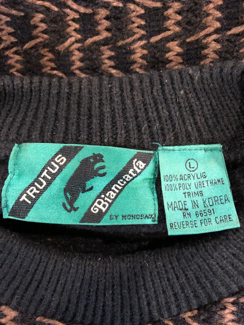 May include: A black and brown sweater with a geometric pattern. The sweater has a green tag with the text "TRUTUS Biancar&iacute;a BY MONOBAIK" and a white tag with the text "100% ACRYLIG 100% POLY URETHANE TRIMS MADE IN KOREA RN 66591 REVERSE FOR CARE".