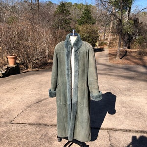 May include: A long, green suede coat with a fur collar and cuffs. The coat is on a mannequin and is standing on a gray concrete surface.