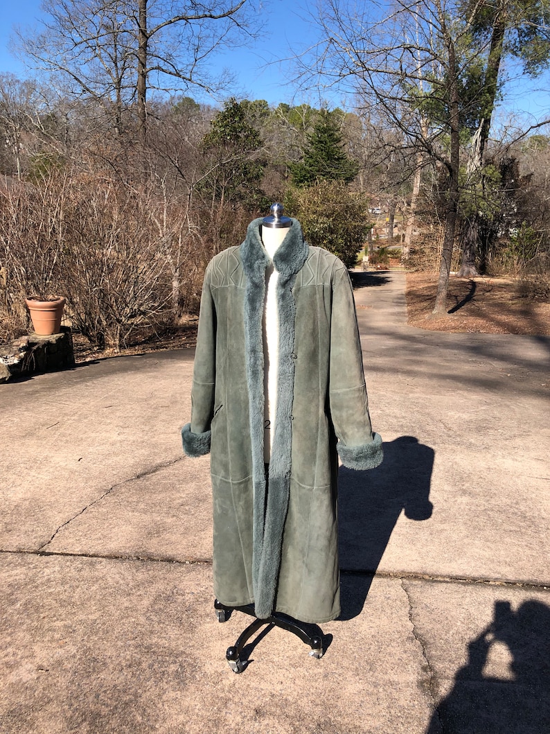 May include: A long, green suede coat with a fur collar and cuffs. The coat is on a mannequin and is standing on a gray concrete surface.