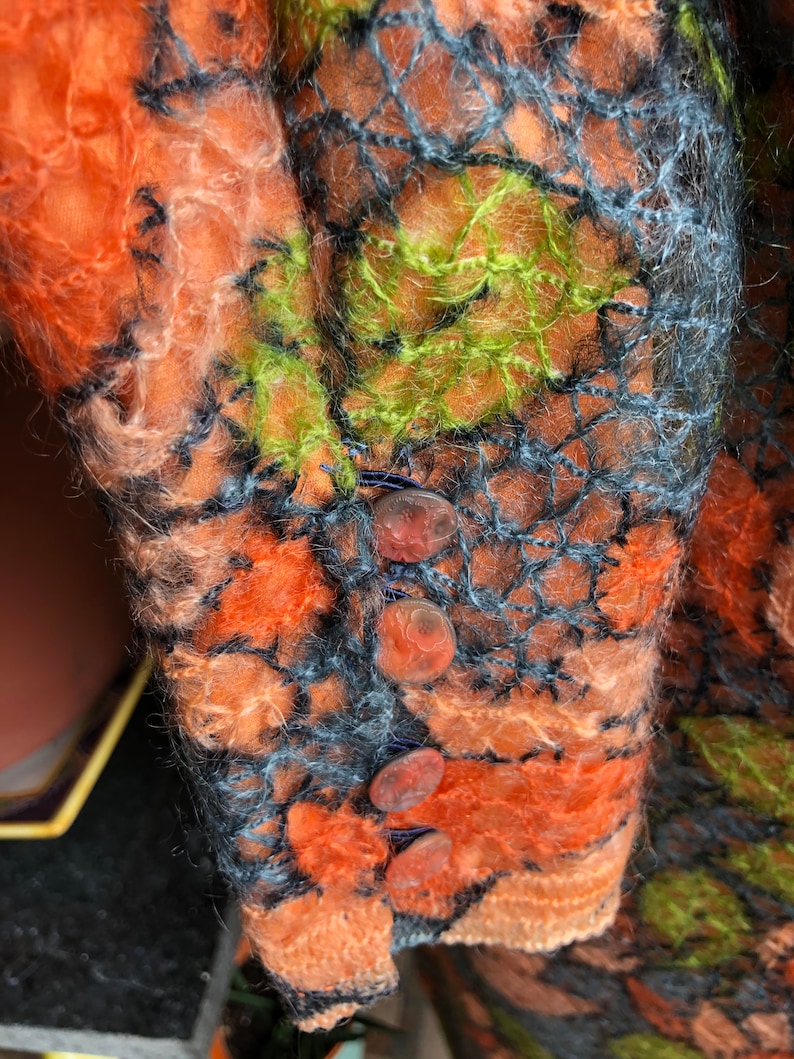Peut inclure: Gros plan d'un v&ecirc;tement tricot&eacute; avec une surface textur&eacute;e et duveteuse. Le v&ecirc;tement est orange, vert et noir avec un motif en filet noir. Il y a quatre gros boutons ronds orange sur le v&ecirc;tement.